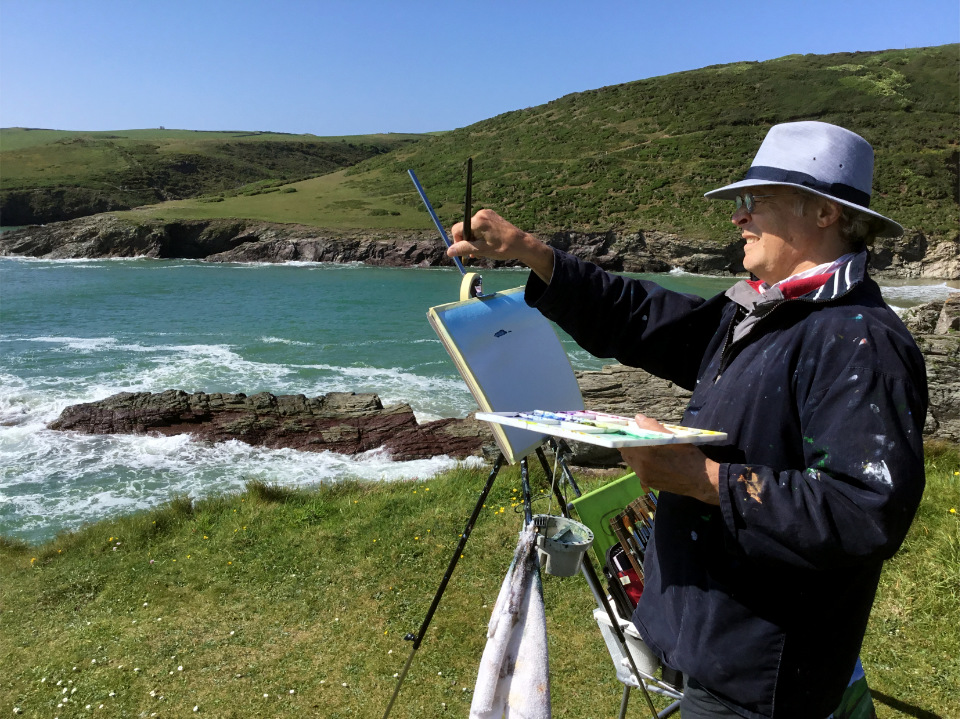 Tony stands at an easel on a grassy clifftop, holding up a brush to gauge proportions while painting a coastal scene. Turquoise waves break over dark rocks in a cove, framed by green headlands and a clear blue sky.